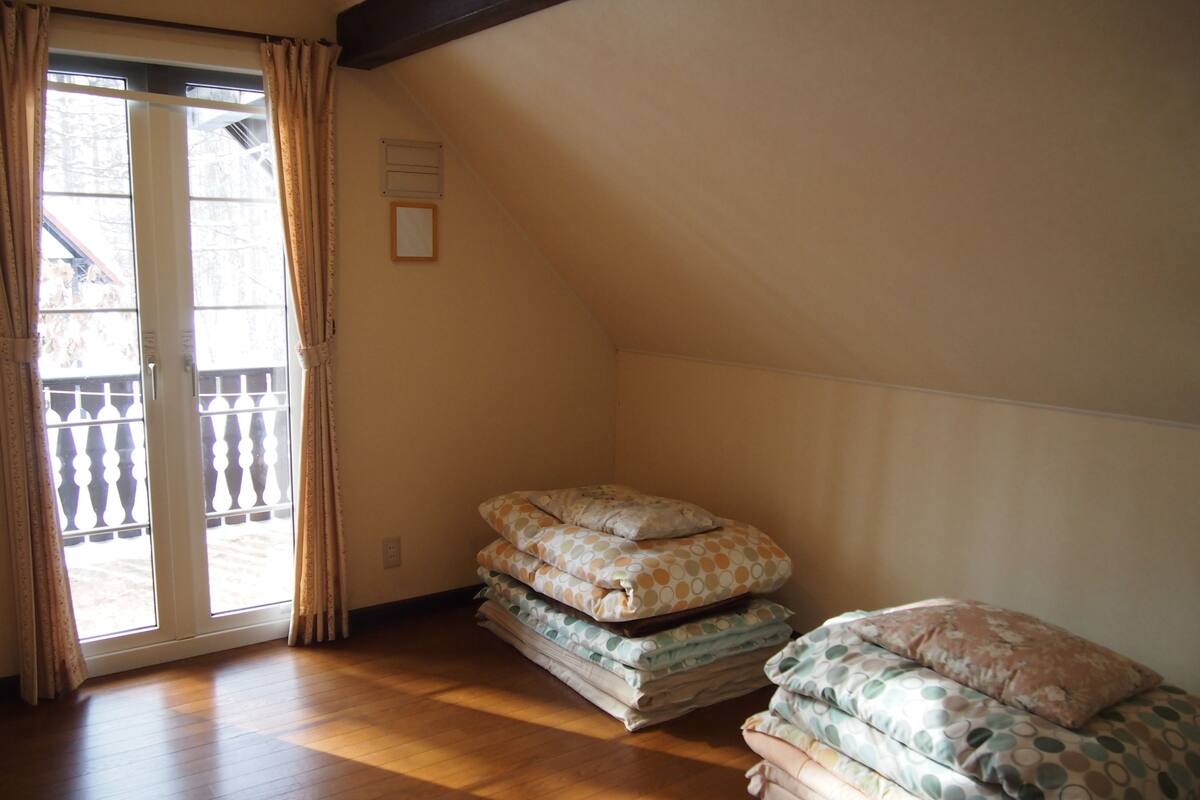 A room with a wooden floor, a sliding glass door with curtains, and a stack of mattresses with patterned bedding.