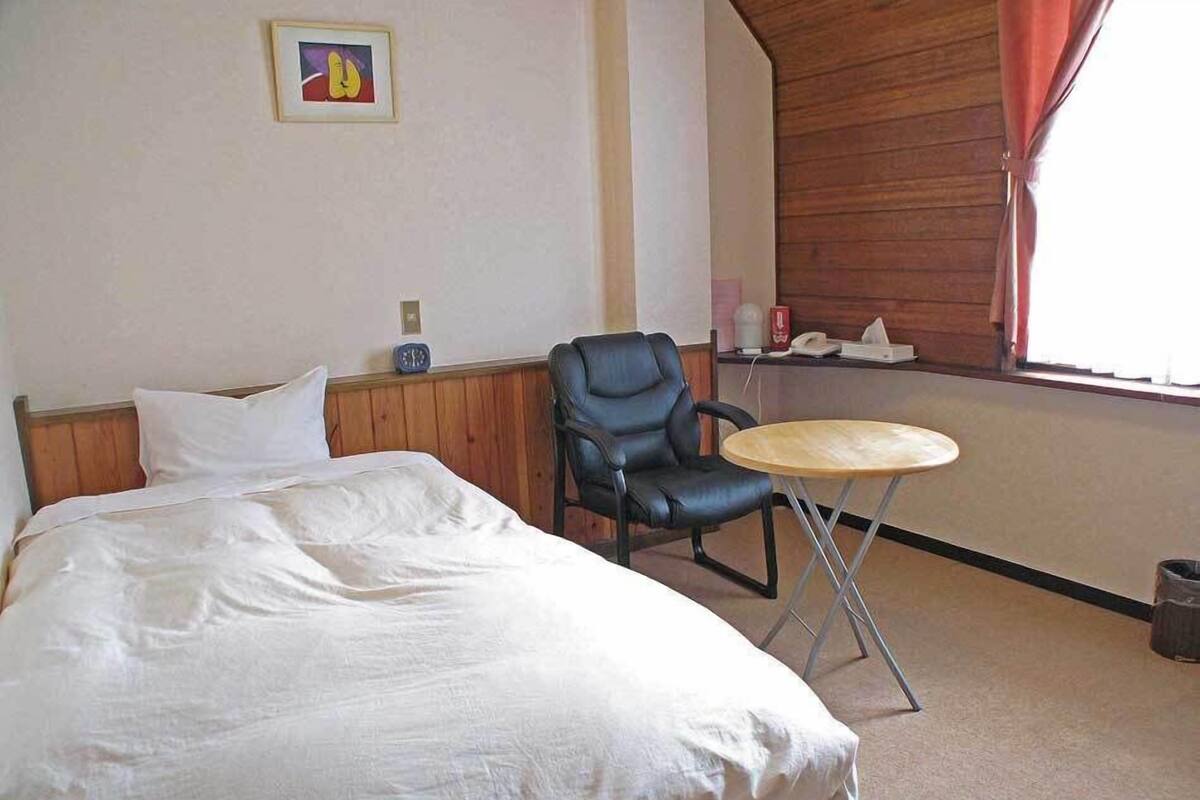Une chambre d’hôtel avec un lit, une chaise, une petite table ronde, une horloge et une fenêtre avec des rideaux.