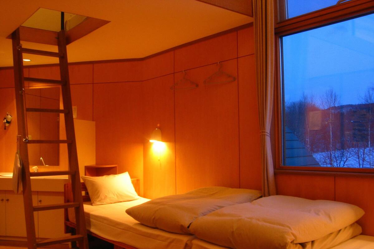 A room with a bunk bed, a ladder, a bedside table, a hanging light, and a window with a view of trees and buildings.