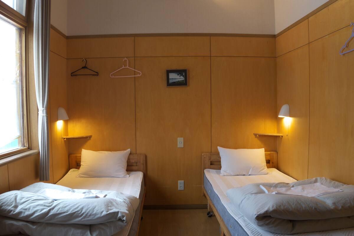 A hotel room with two single beds, wooden panel walls, bedside lamps, and a window with curtains.