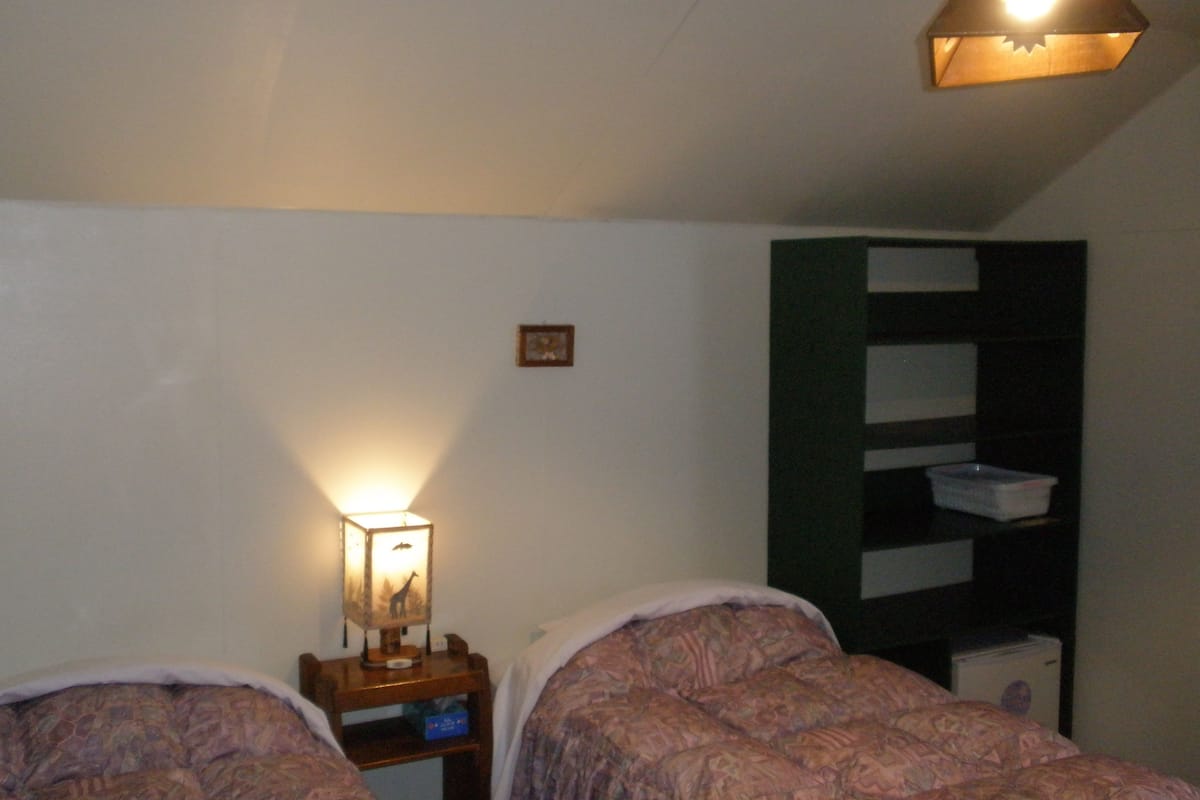 Quarto com duas camas, mesa de cabeceira com abajur, prateleira pequena e prateleira na parede.