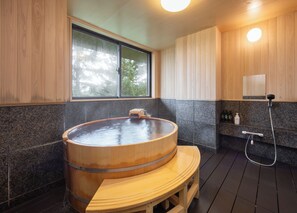 Una bañera de madera en una habitación con ventana, ducha y estante con artículos de aseo.