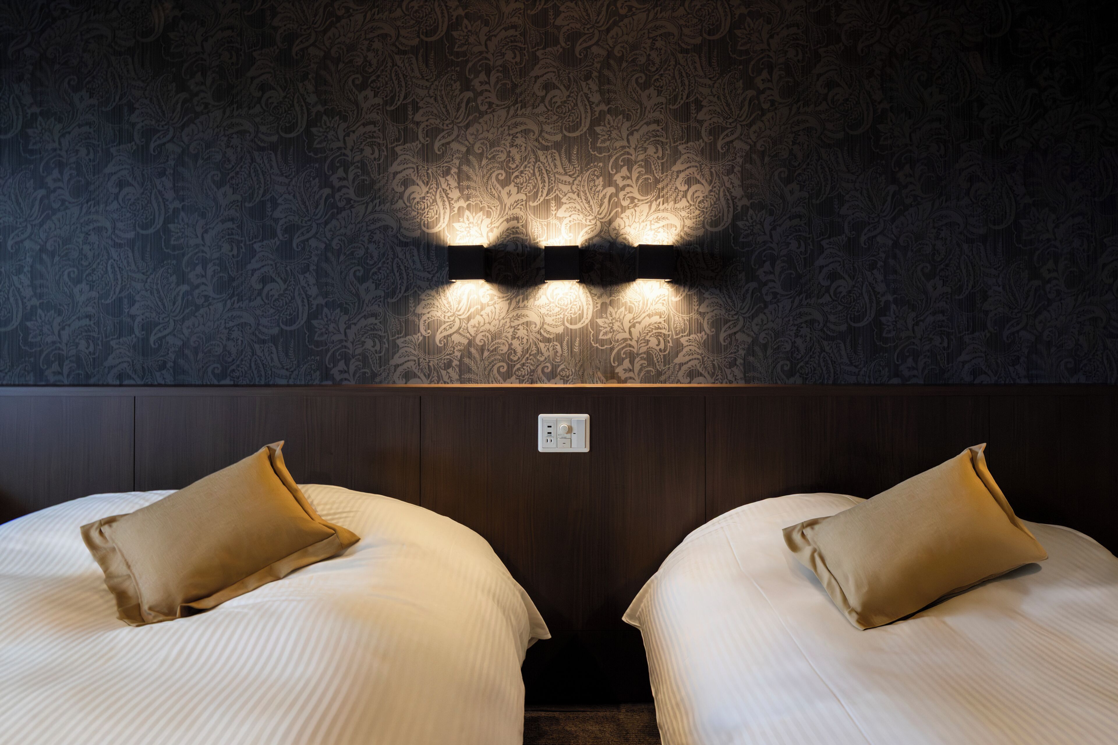 Two beds with beige pillows, a patterned wallpaper, and wall-mounted lighting.