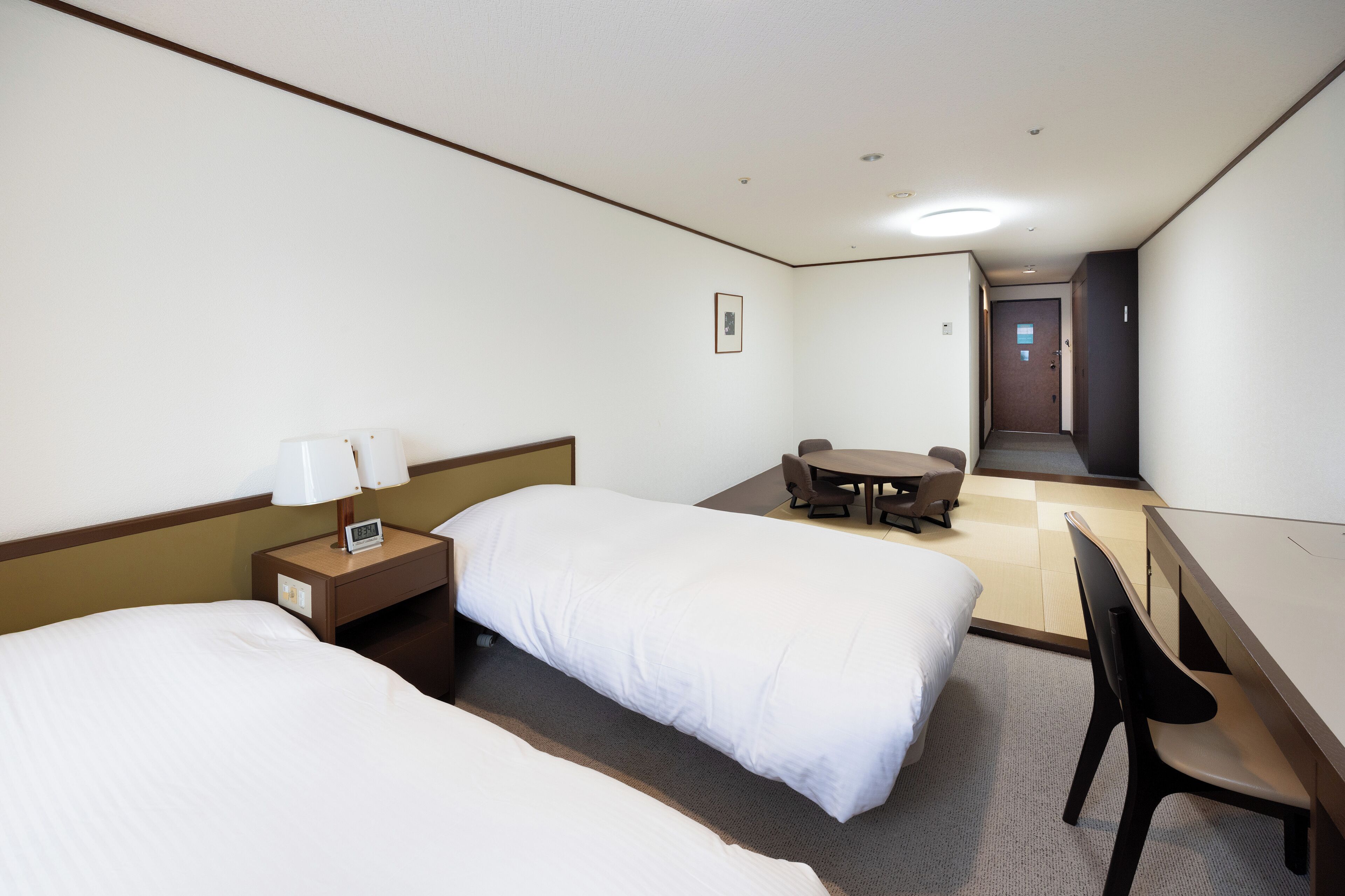 A hotel room with two beds, a desk, a chair, a small table, and a door leading to another room.