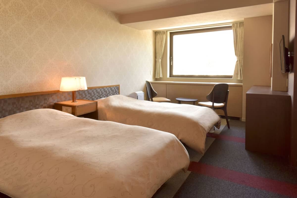 A hotel room with two beds, a desk, a chair, a lamp, and a window with curtains.