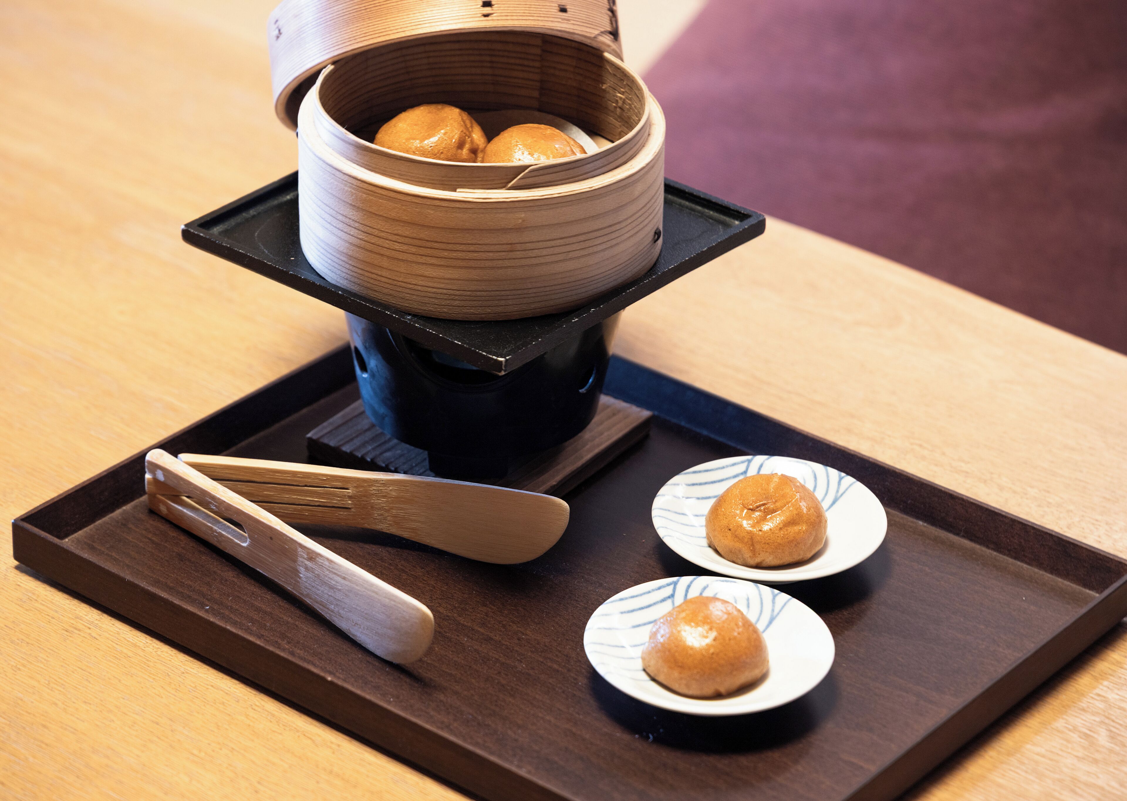 A bamboo steamer with two dumplings on a tray, accompanied by chopsticks and a small plate with a single dumpling.