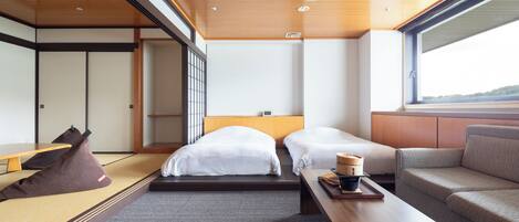 Quarto de hotel moderno com teto de madeira, duas camas com roupa de cama branca, um sofá, uma pequena mesa com bandeja e vista através de uma grande janela.