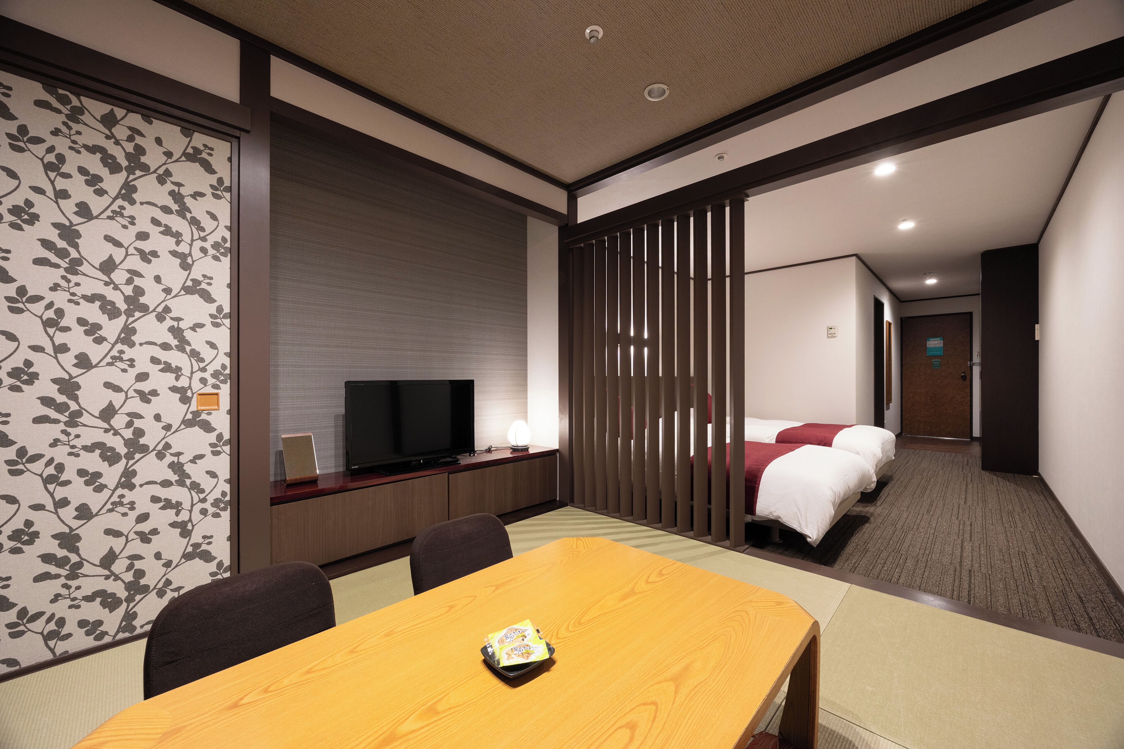 A hotel room with a wooden dining table, a TV, a bed, and a sliding door.