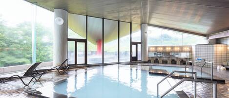 An indoor swimming pool with clear glass walls, a tiled floor, and a bar area with seating.