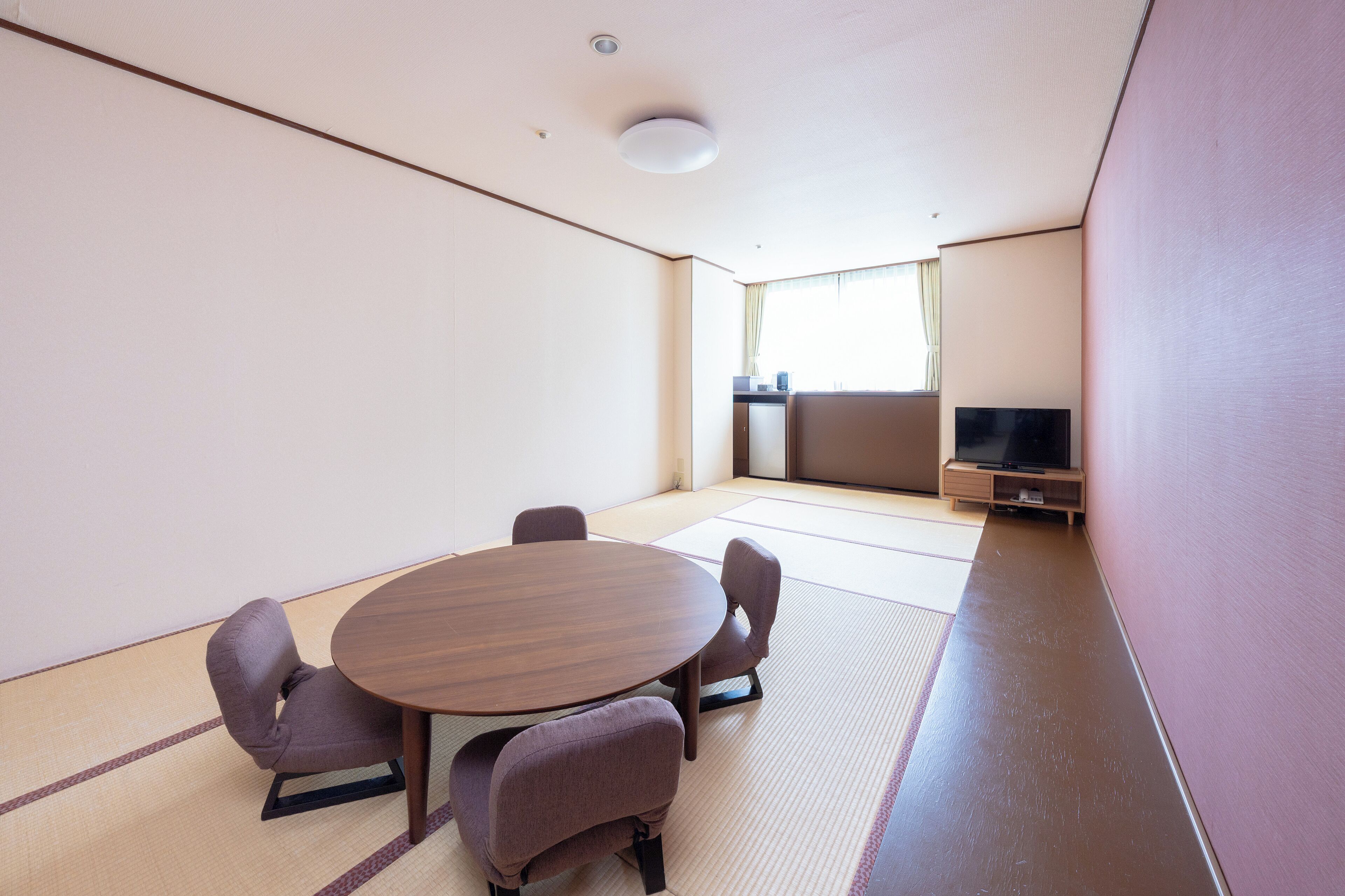 Una habitación con suelo de tatami, una mesa redonda, cuatro sillas, un televisor sobre un soporte y una ventana con cortinas.
