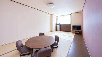 Una habitación con suelo de tatami, una mesa redonda, cuatro sillas, un televisor sobre un soporte y una ventana con cortinas.