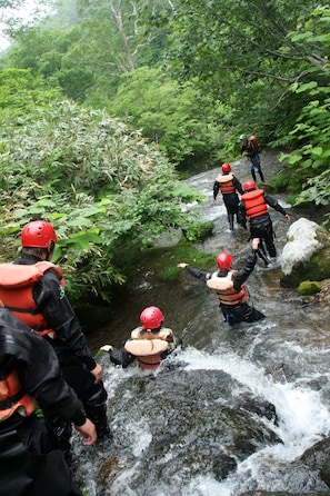 A group of people wearing life jackets and helmets navigating a rocky stream in a forested area.