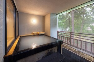 Un jacuzzi con borde de madera, una luz empotrada en la pared y vistas a árboles a través de un balcón cubierto.