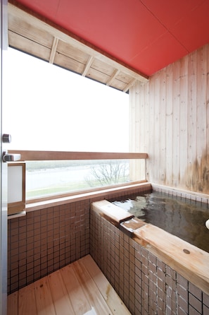 A wooden bathhouse with a tiled floor, a wooden bench, and a window overlooking a body of water.