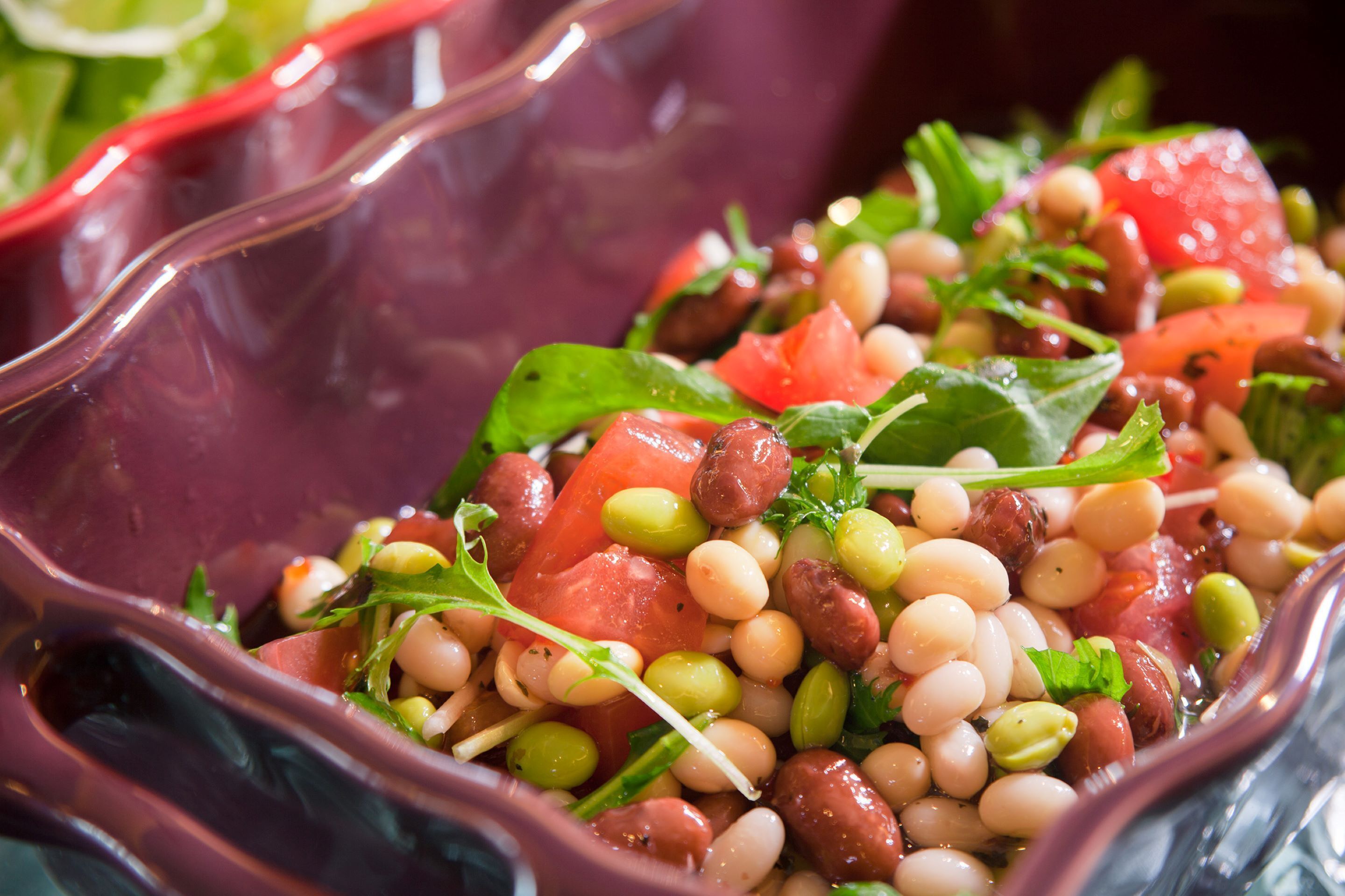 Close-up van een kleurrijke salade met verschillende bonen, tomaten en groene bladgroenten in een glazen schaal.