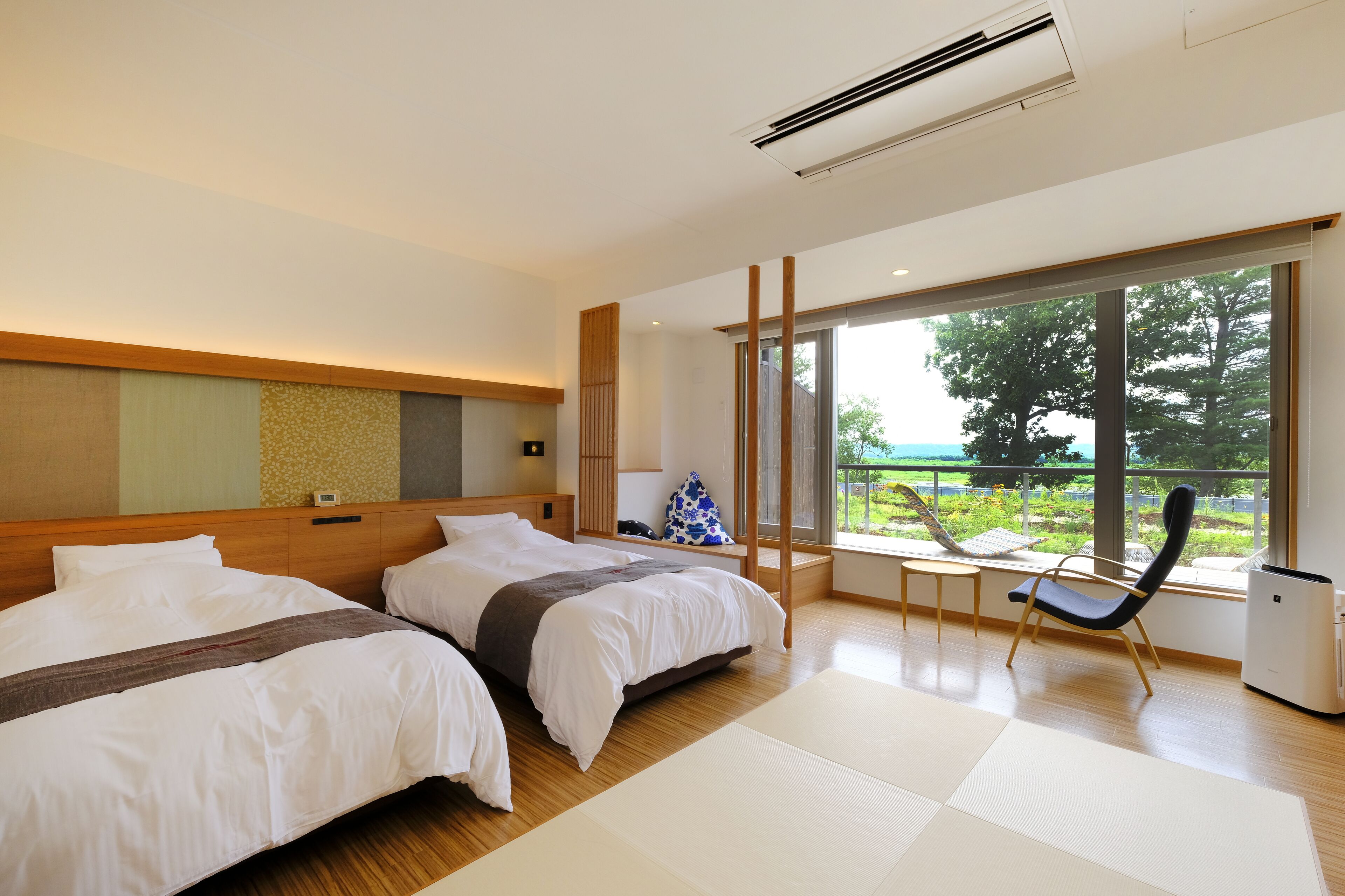 A modern hotel room with two beds, a small table, a chair, and a large window offering a view of greenery.