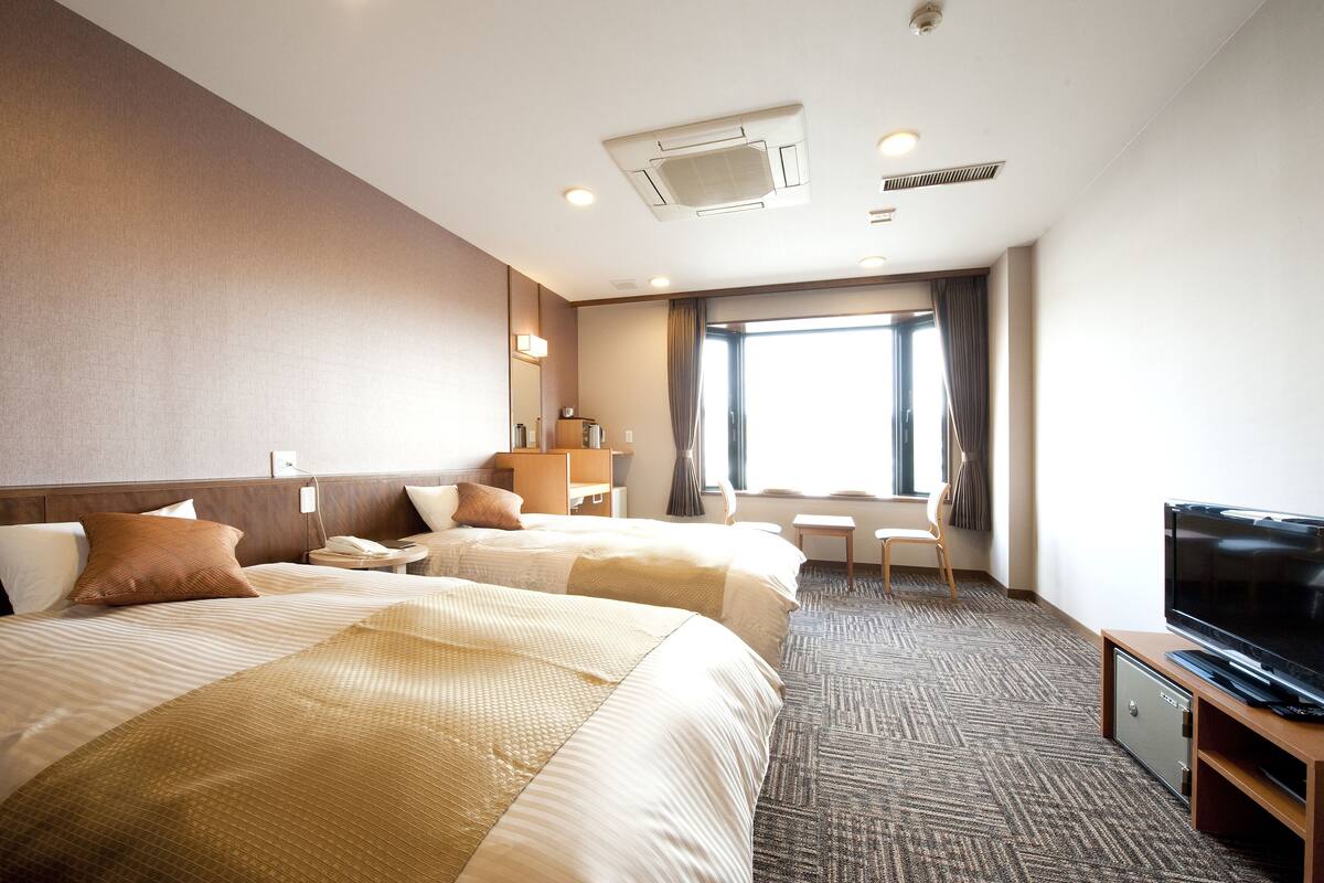 A hotel room with two beds, a TV, a desk, and a window with curtains.
