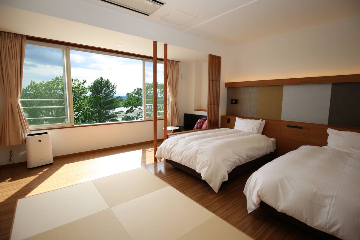 A hotel room with two beds, a large window with curtains, a wooden floor, and a view of trees and buildings outside.