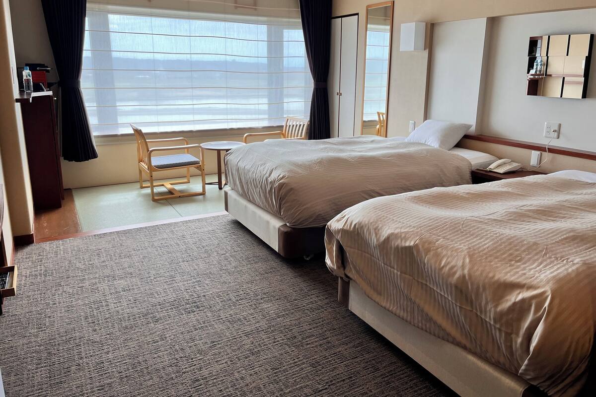 A hotel room with two beds, a small table, a chair, a mirror, and a window with curtains.