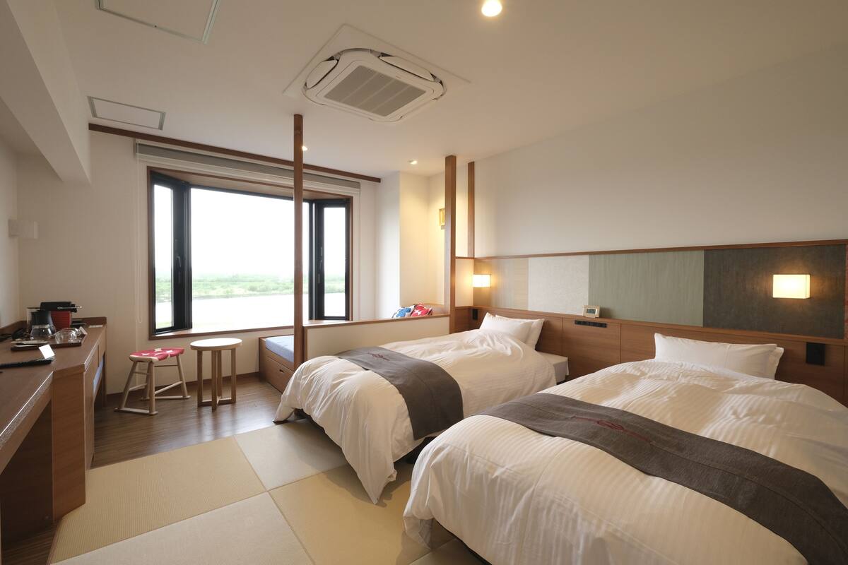A hotel room with two beds, a desk with a chair, a small stool, and a window with a view.