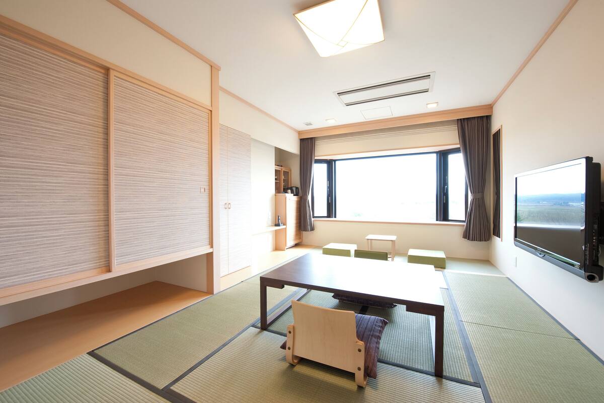 A room with tatami flooring, a low table, a chair, a television, and a large window with curtains.