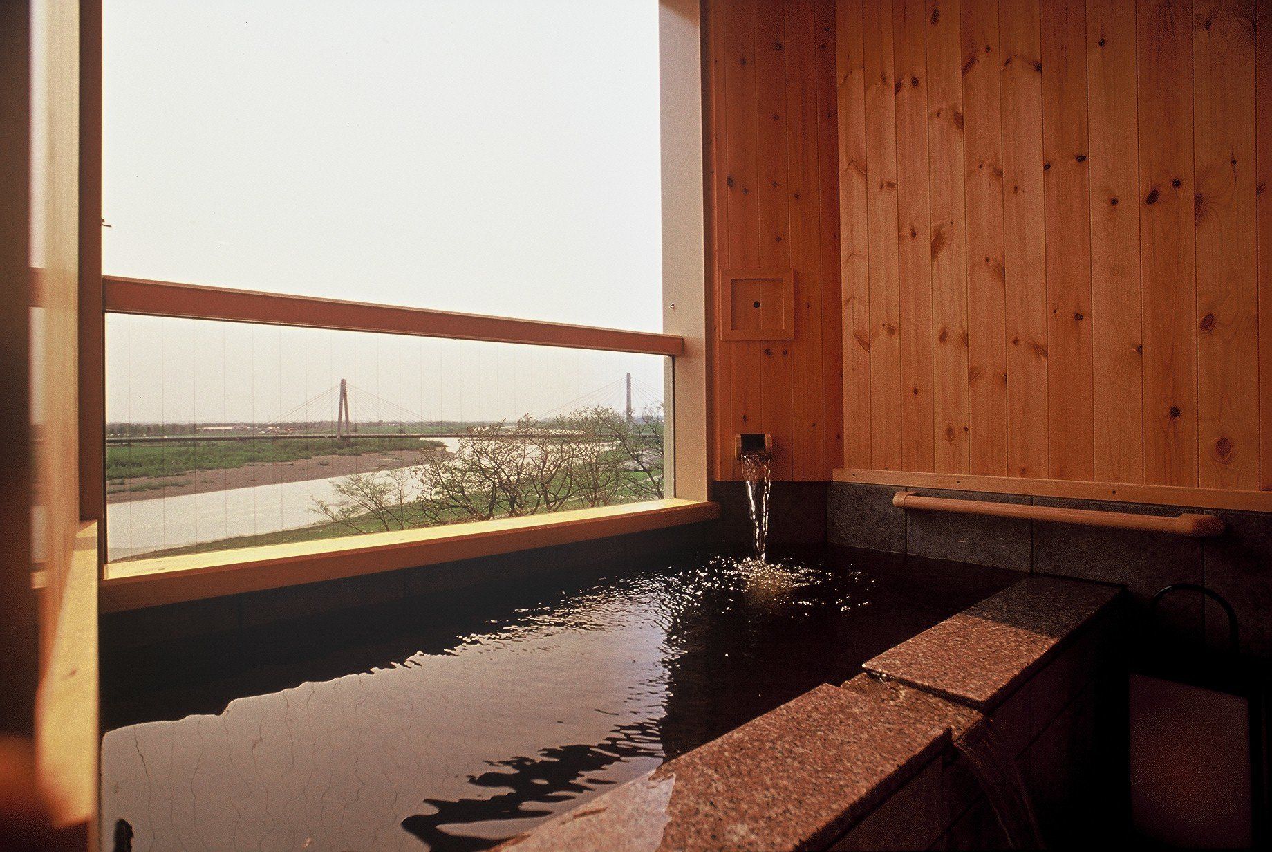 A wooden cabin with a large window overlooking a river and a bridge.