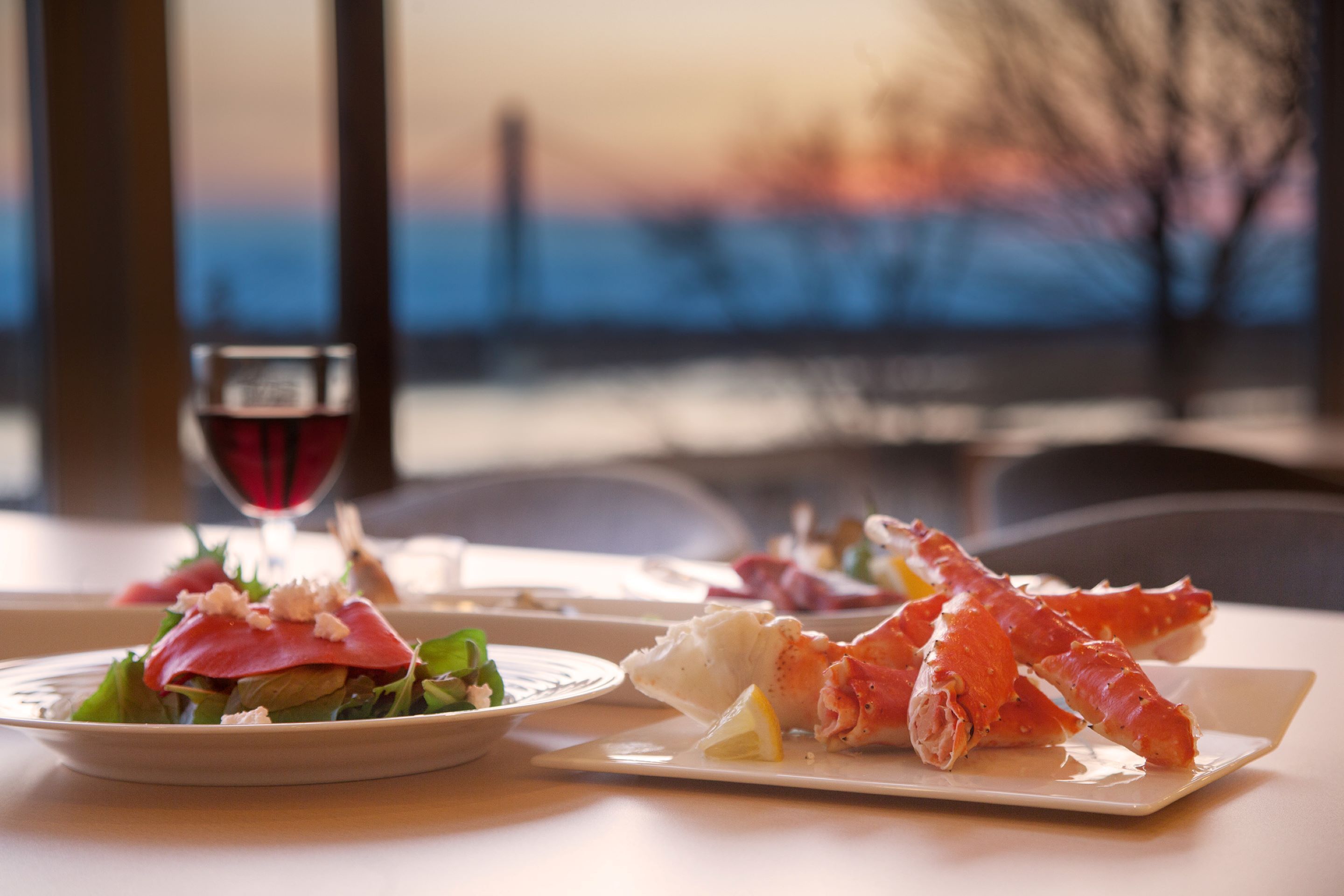A table with plates of food, a glass of red wine, and a view of the sea.