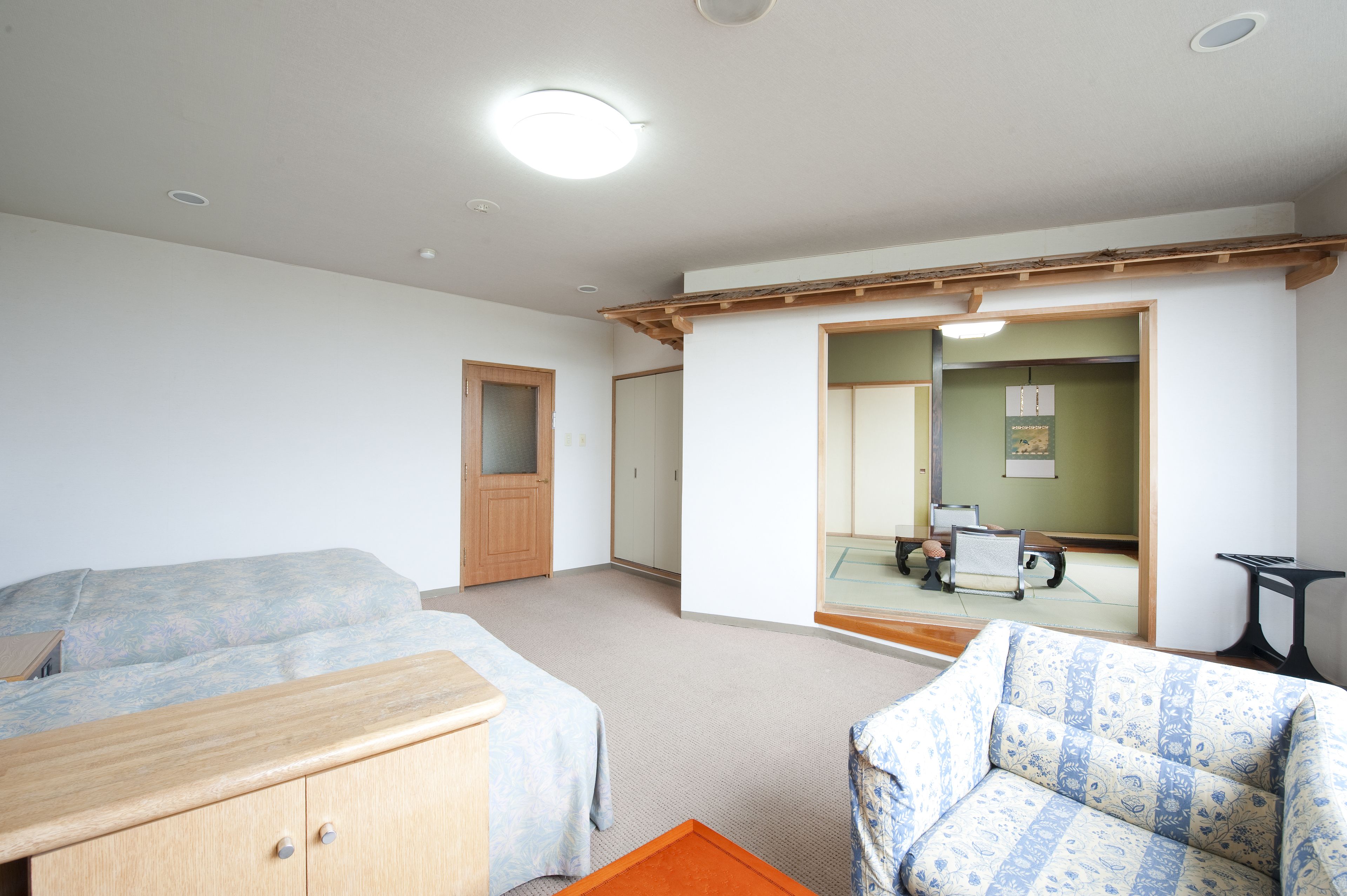 A room with two beds, a wooden dresser, a sofa, and a mirror reflecting a traditional Japanese room with tatami flooring and a shoji screen.