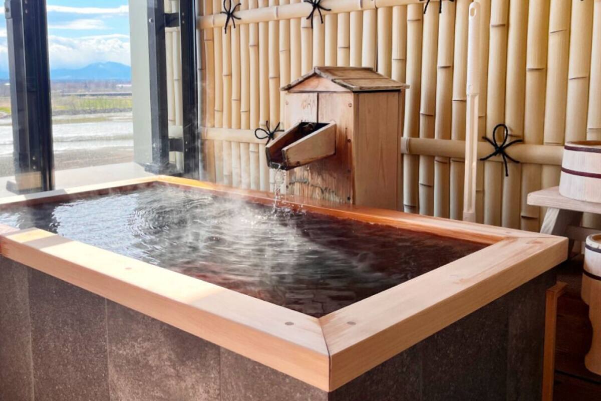 A hot spring bath with a wooden edge, bamboo wall, and a wooden structure with a water spout.