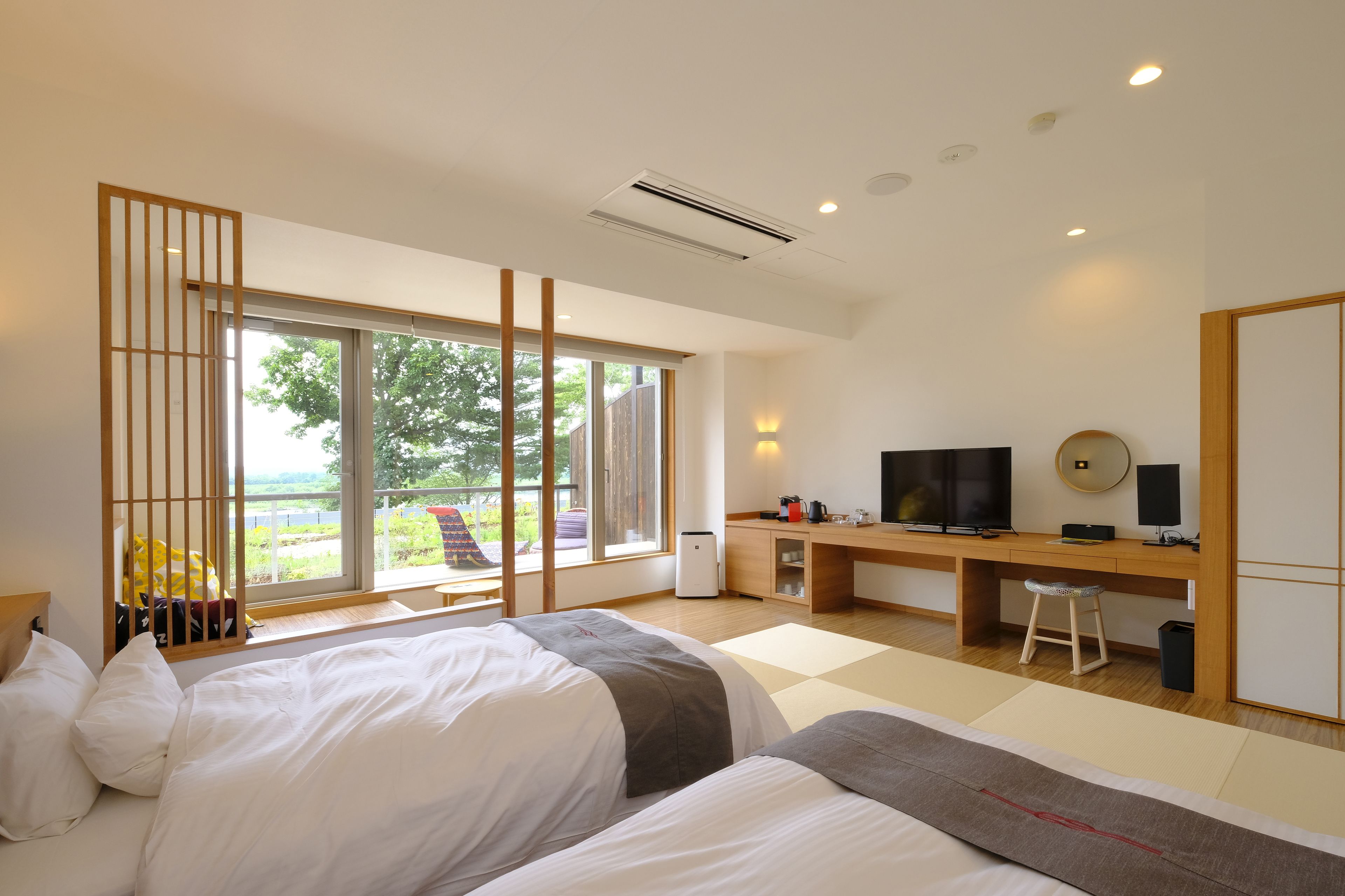A modern hotel room with two beds, a desk, a TV, and a large window offering a view of the outdoors.