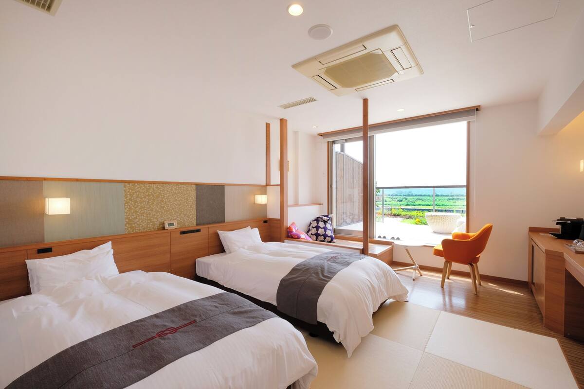 A hotel room with two beds, a desk, a chair, and a large window offering a view.