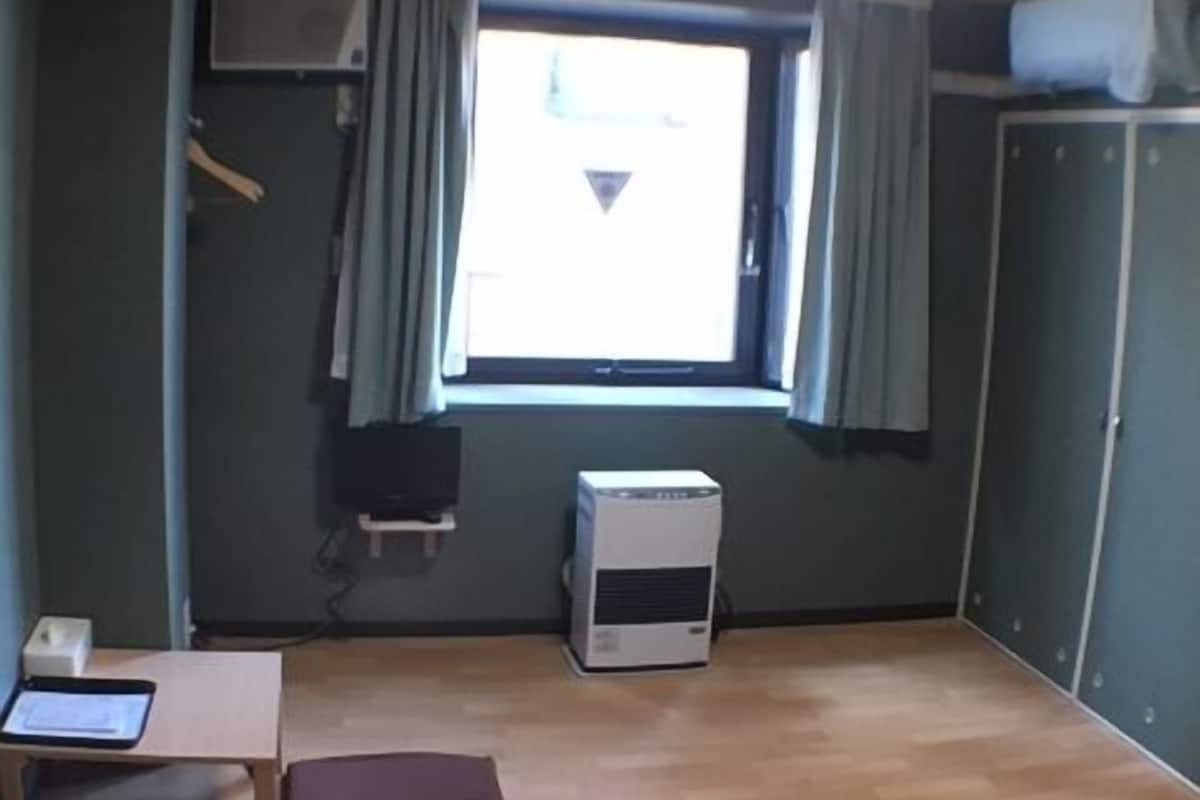 Um quarto com janela, radiador, escrivaninha e cadeira, e guarda-roupa.