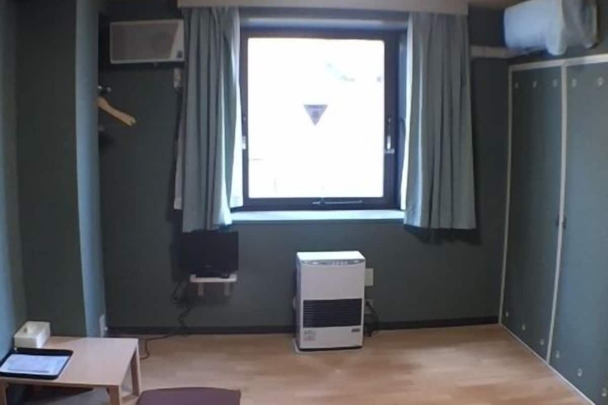 Um quarto com piso de madeira, uma janela com cortinas, uma pequena mesa com um tablet, uma almofada vermelha e um aparelho de ar condicionado branco.