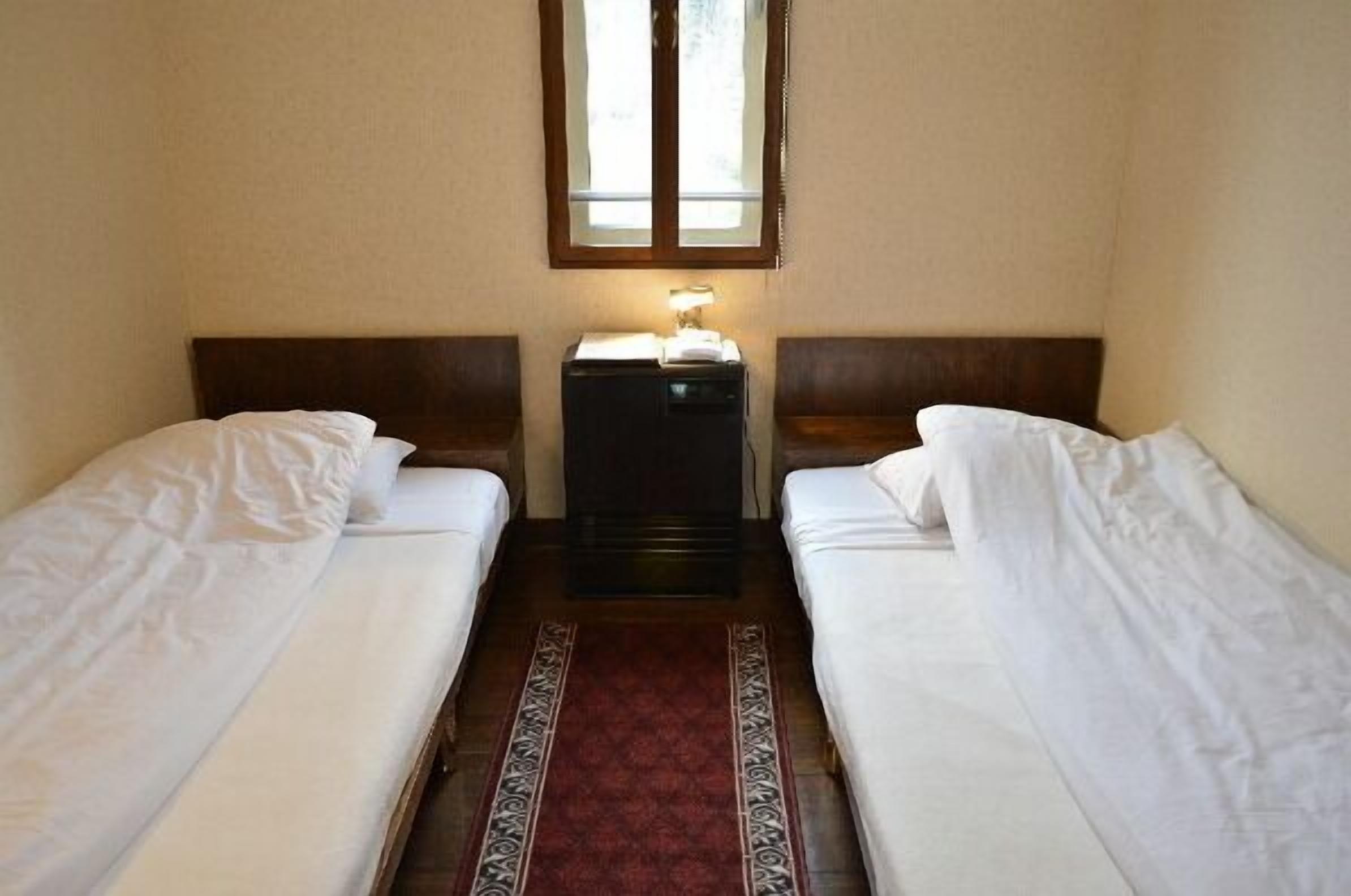 A small, simple room with two single beds, a nightstand with a lamp, and a window above the beds.