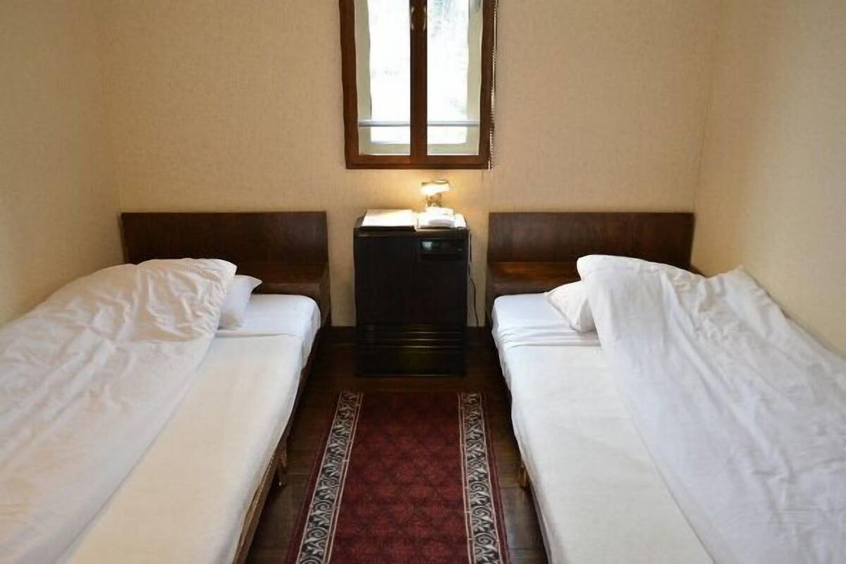 A small, simple room with two single beds, a nightstand with a lamp, and a window above the beds.