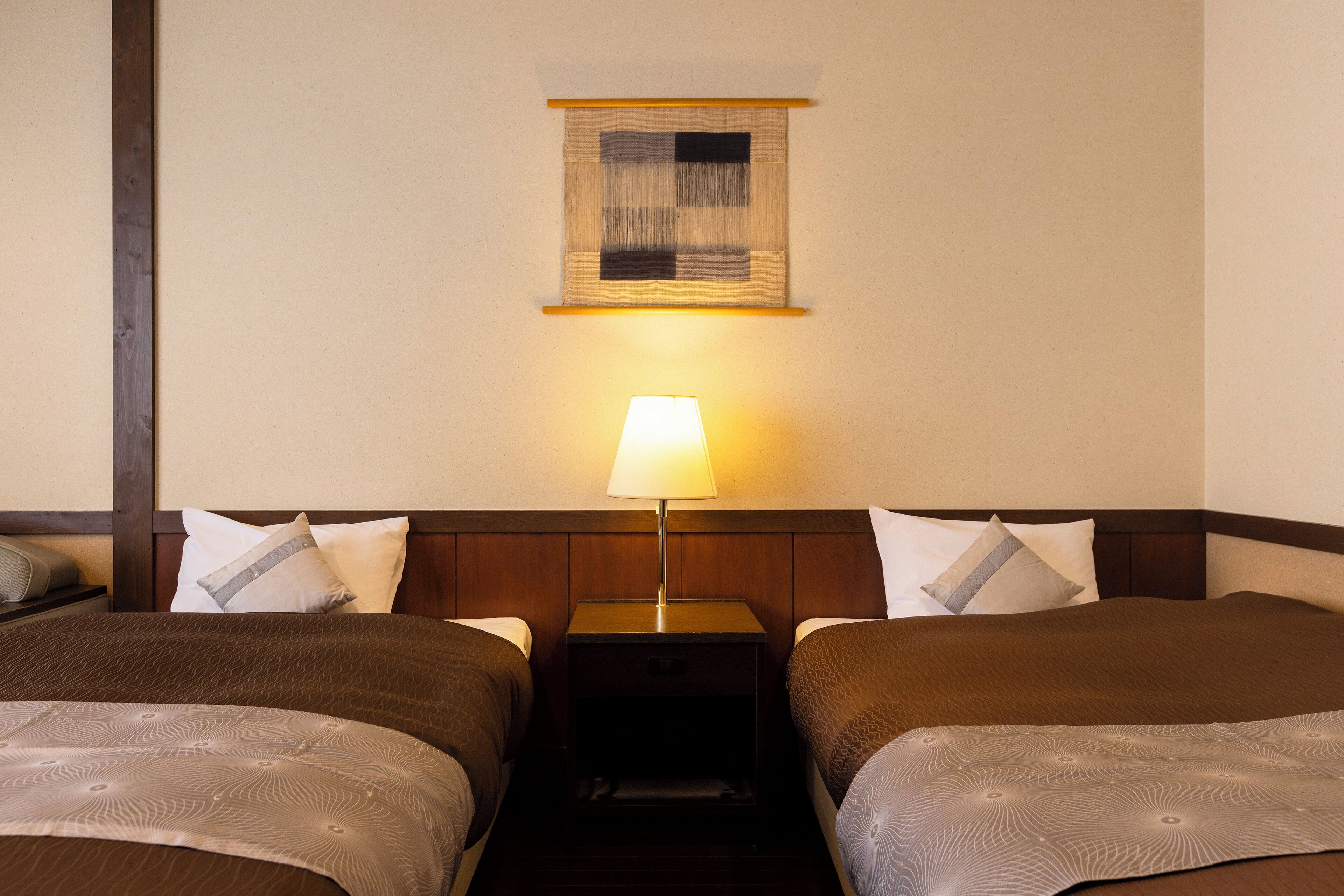 Hotellihuone, jossa on kaksi sänkyä, yöpöytä lampun kanssa ja pieni seinälle kiinnitetty taideteos.