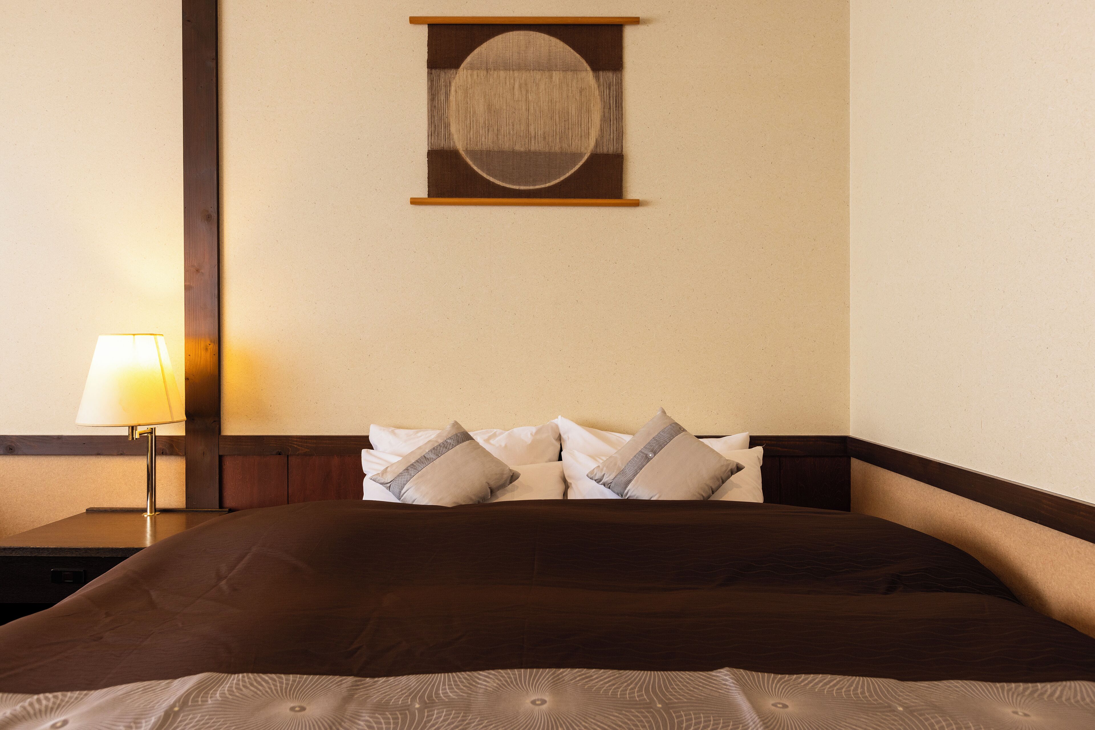 Cama bem arrumada com cabeceira marrom escura, abajur e uma obra de arte circular fixada na parede.