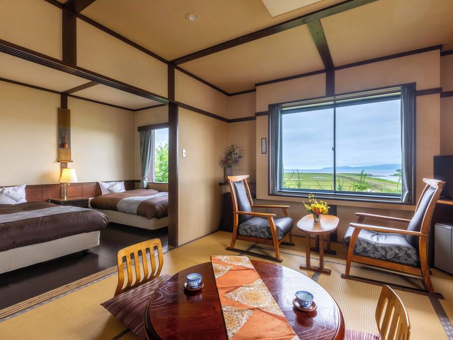 Quarto de hotel com cama, duas cadeiras, uma mesinha e uma janela grande com vista para o exterior.