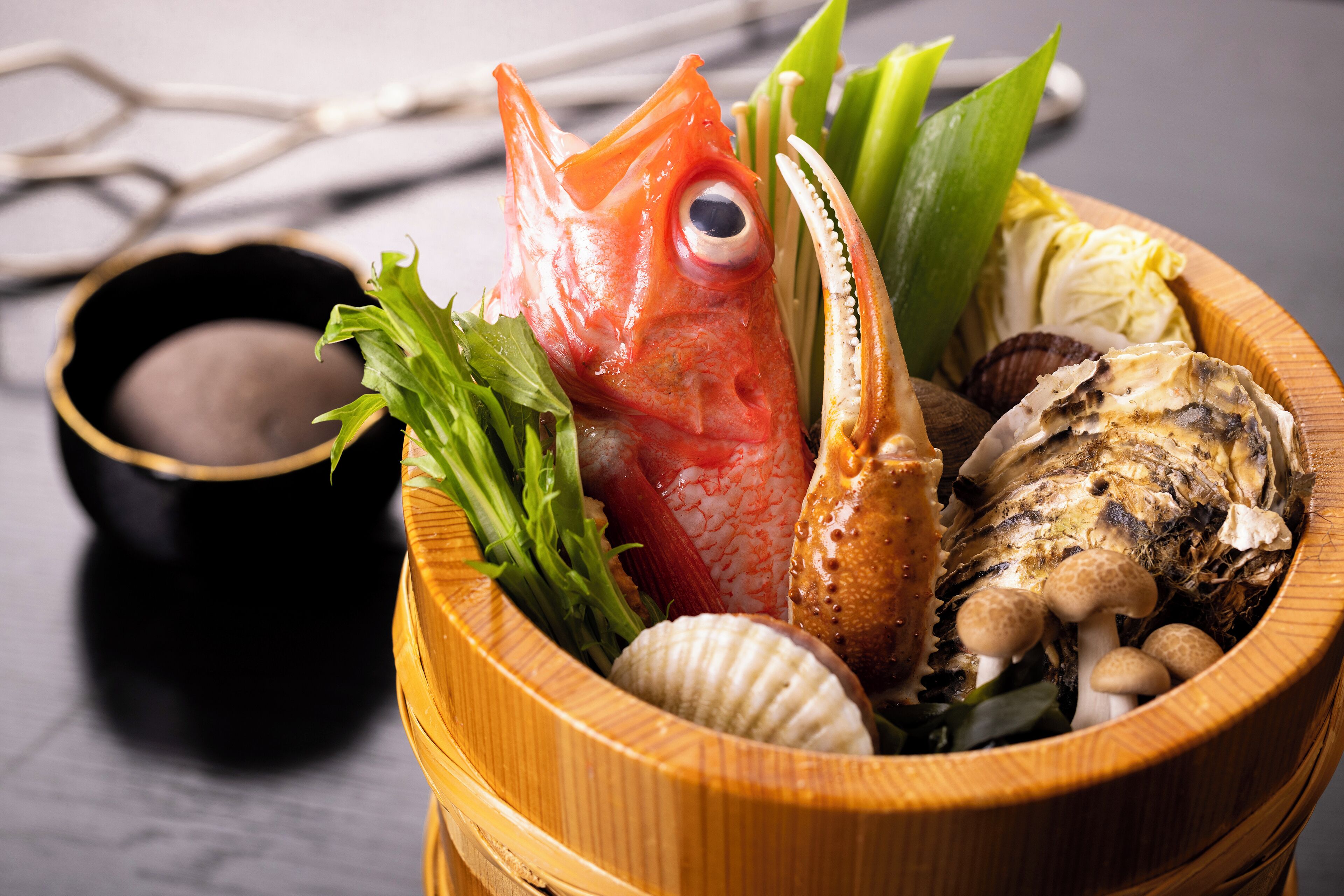 A wooden bowl containing a fish, shellfish, and greens, with a small bowl of sauce beside it.