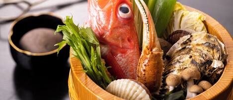 A wooden bowl containing a fish, shellfish, and greens, with a small bowl of sauce beside it.