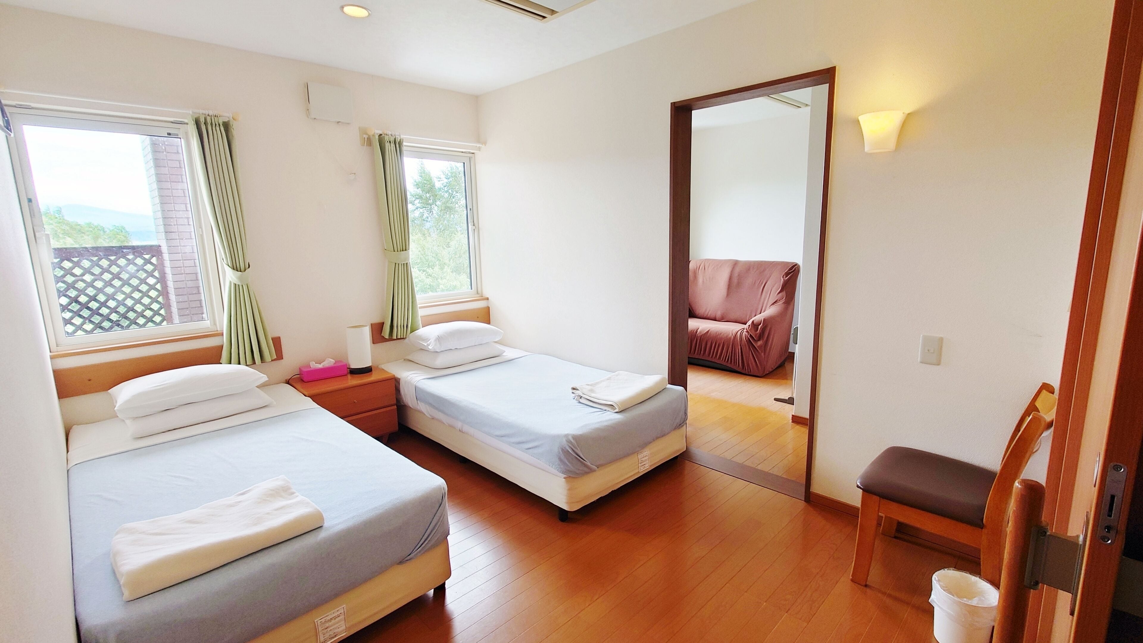 A room with two beds, a wooden floor, a mirror, a sofa, and a window with curtains.