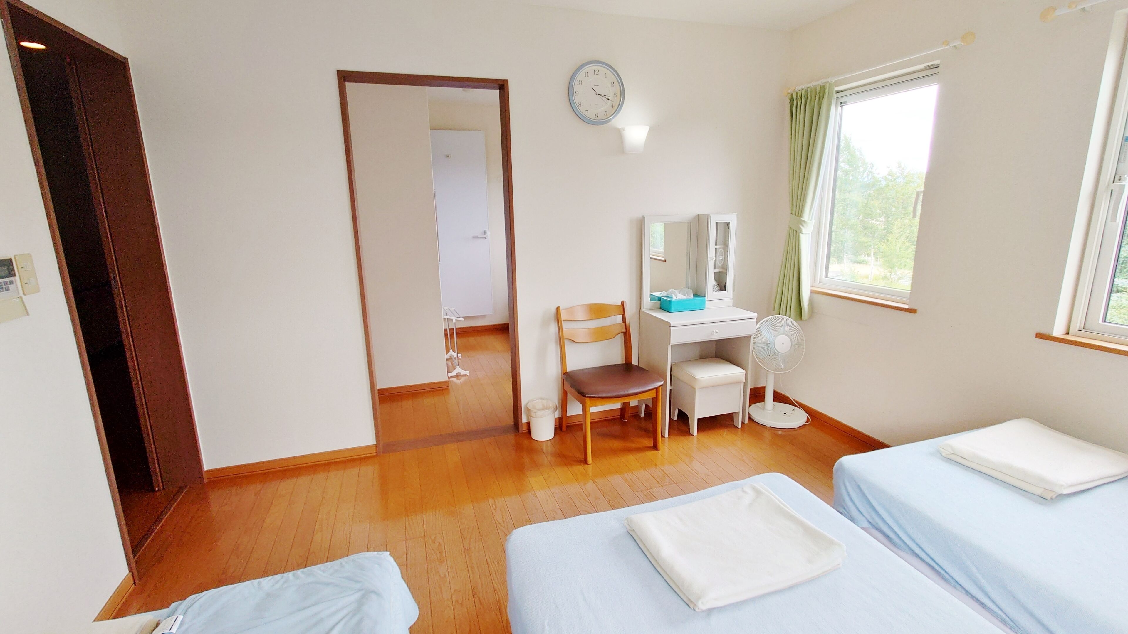 A room with two beds, a desk with a chair, a clock on the wall, and a window with curtains.
