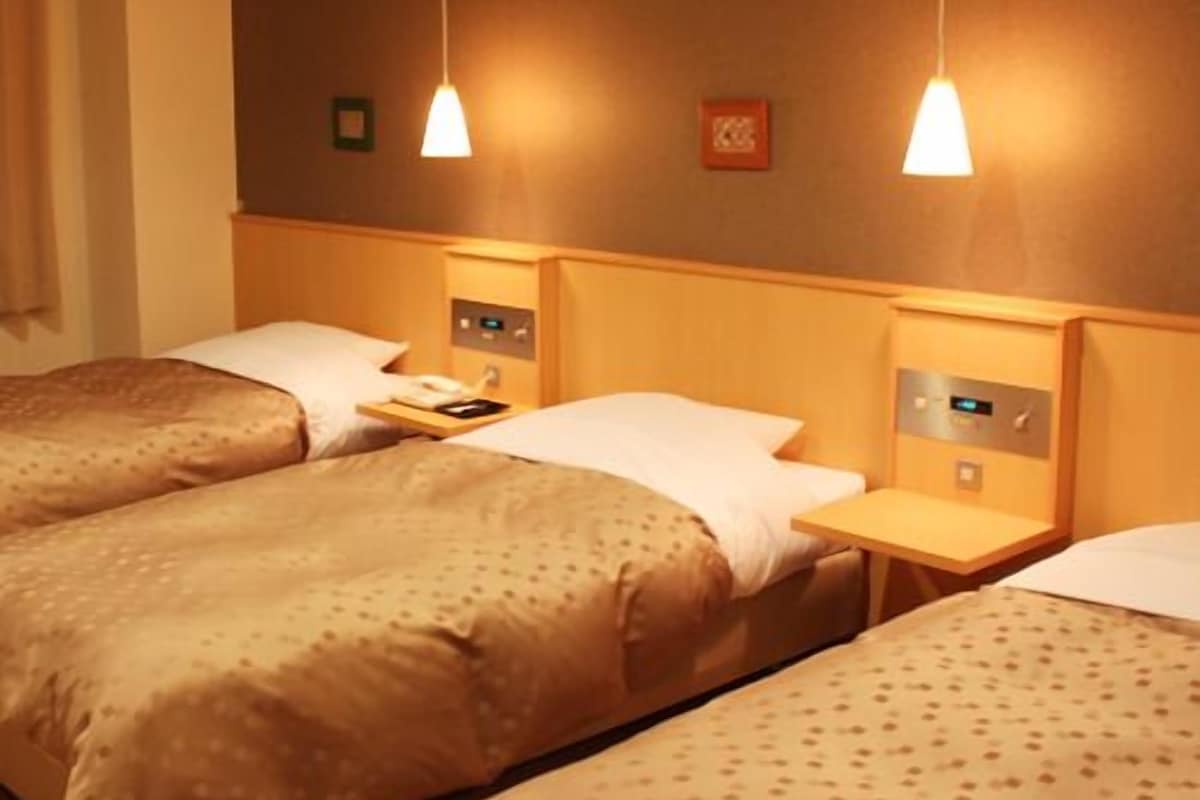 Habitación de hotel con tres camas, cada una con cabecero de madera, una mesita de noche con lámpara y un reloj pequeño.