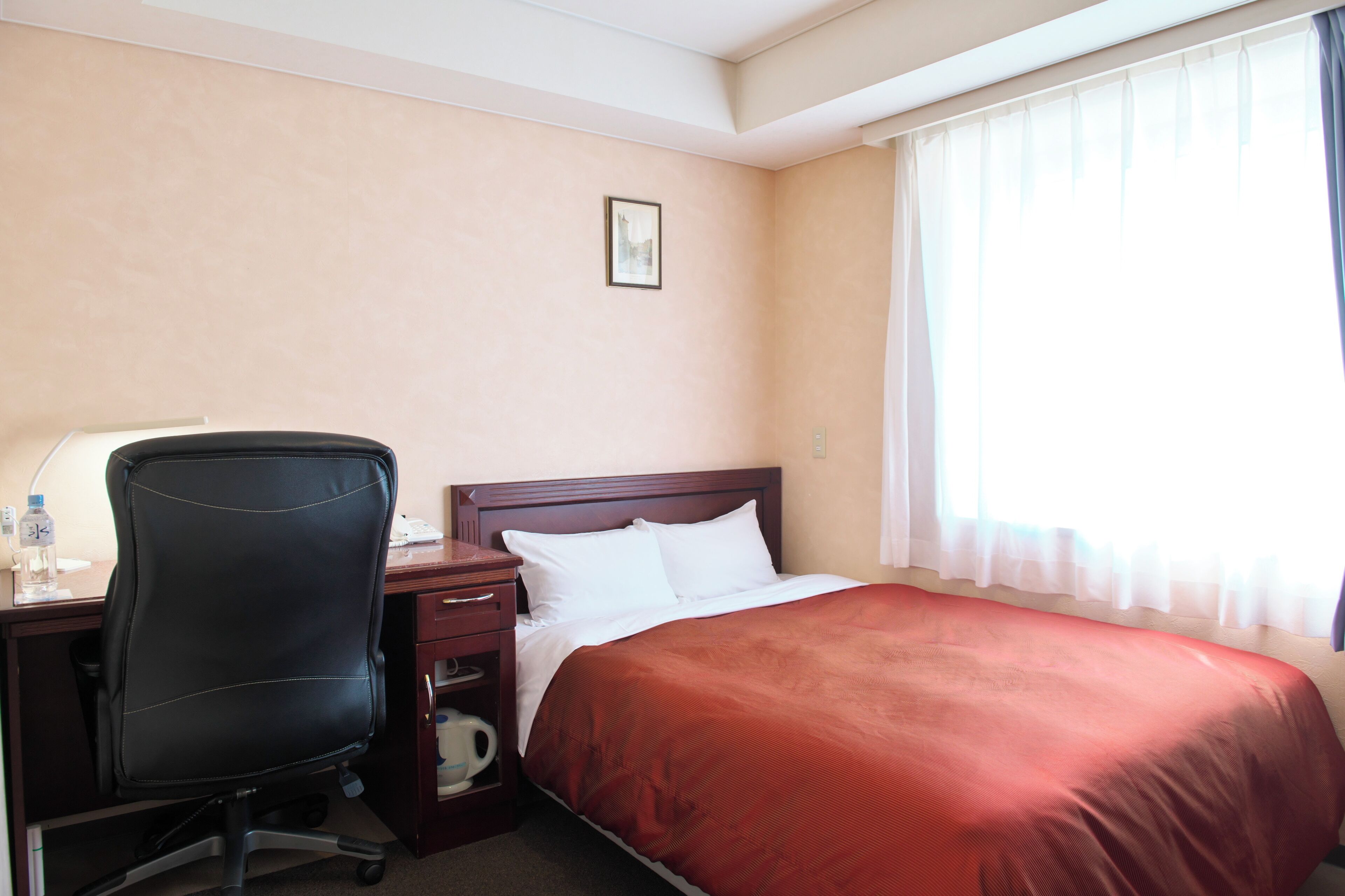 A hotel room with a bed, a desk with a chair, a nightstand, and a window with curtains.
