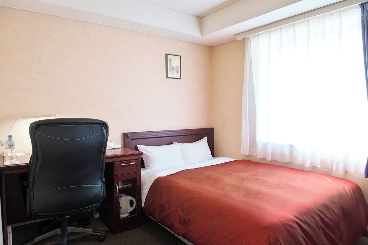 A hotel room with a bed, a desk with a chair, a nightstand, and a window with curtains.