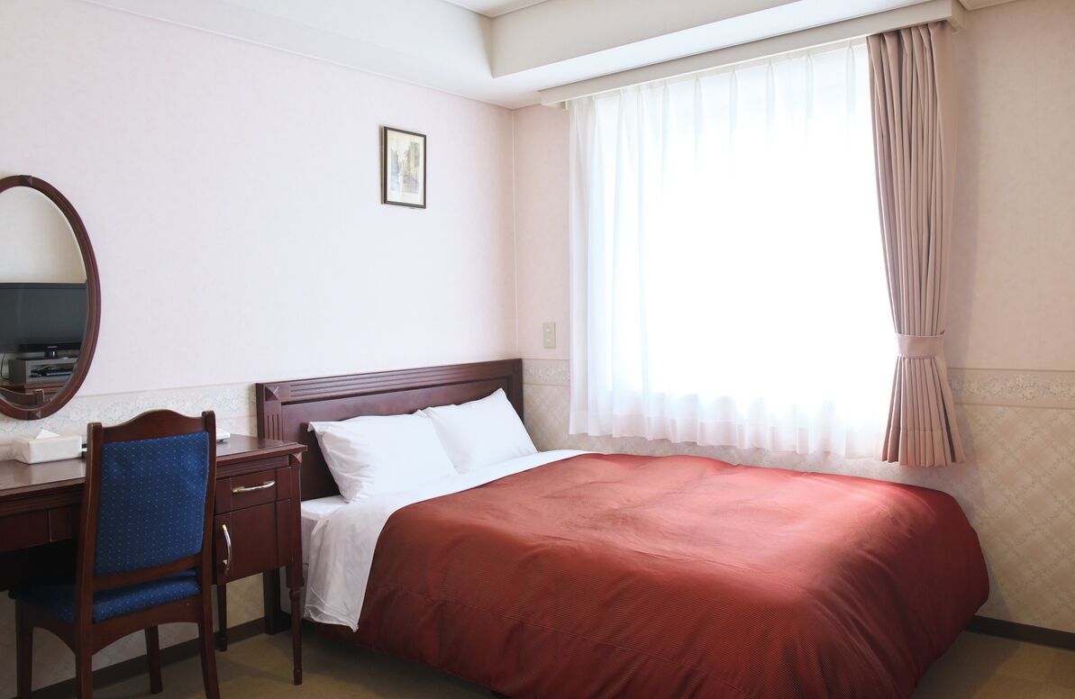 Een hotelkamer met een bed, een bureau met stoel, een spiegel en een raam met gordijnen.