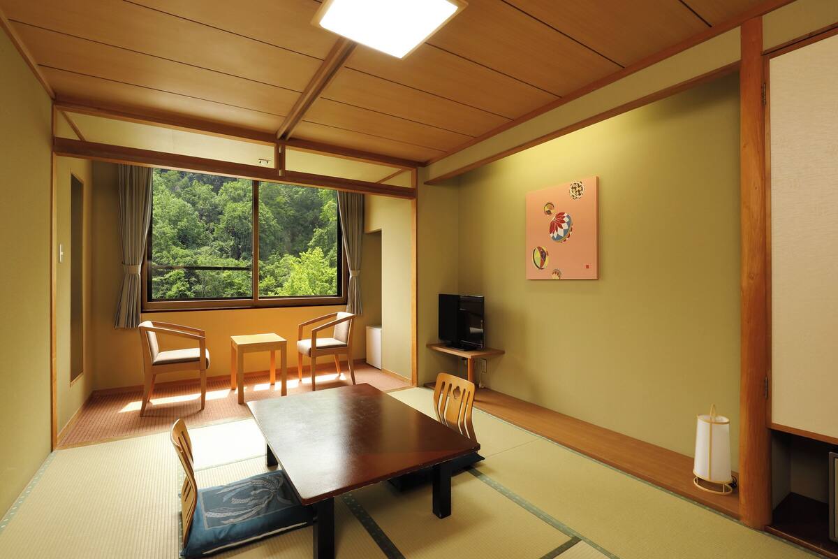 Ett traditionellt japanskt rum med tatamimattor på golvet, ett lågt bord, stolar, en tv och ett fönster med utsikt över träd.