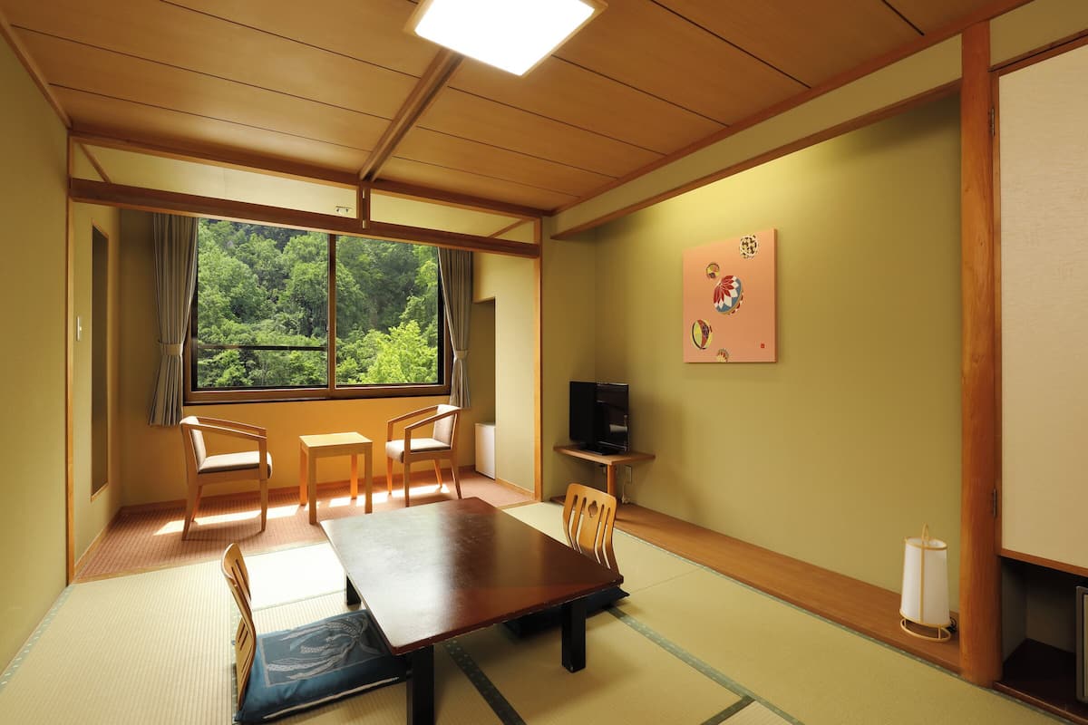 Una tradizionale stanza giapponese con pavimento in tatami, un tavolino, sedie, una televisione e una finestra con vista sugli alberi.