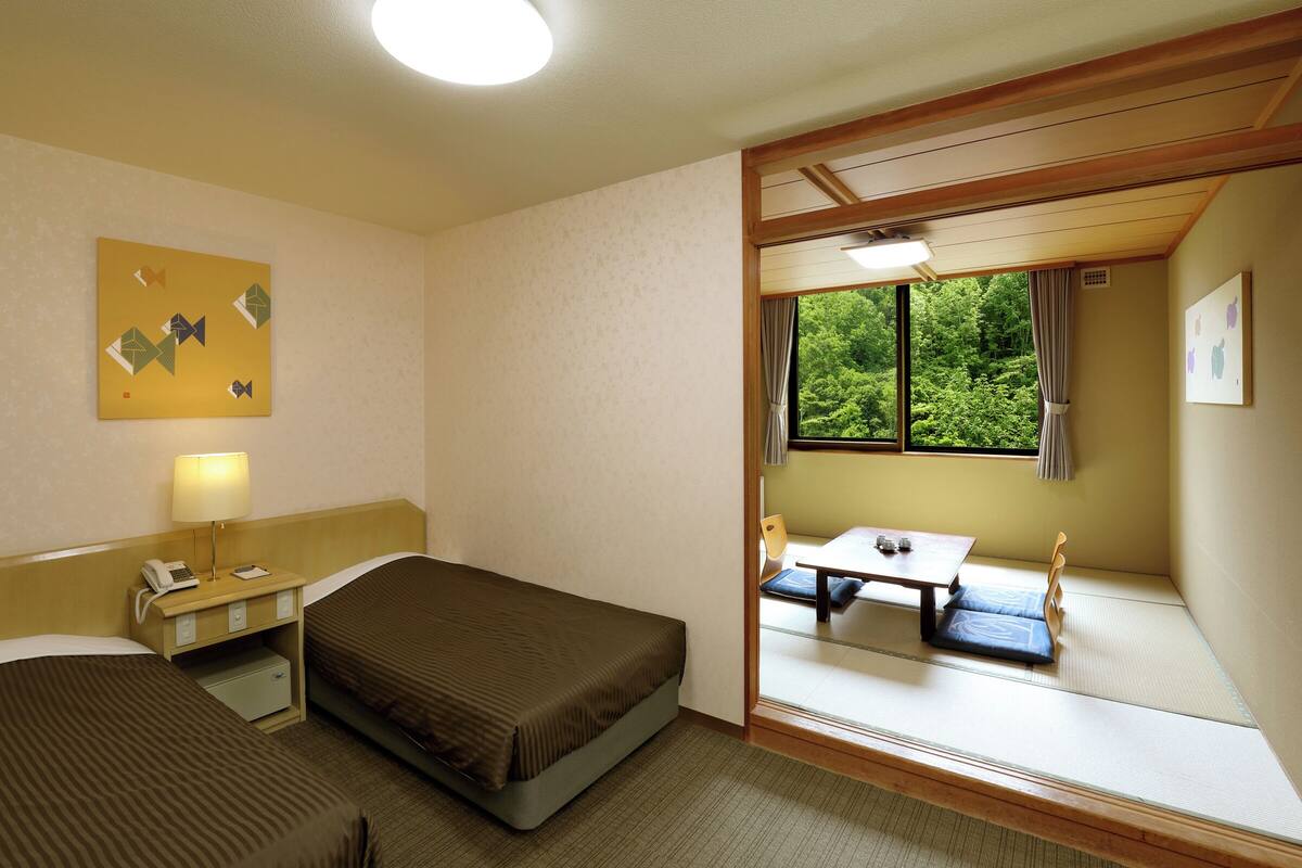 Ett hotellrum med en säng, ett nattduksbord, en lampa och utsikt över en matsal genom ett fönster.