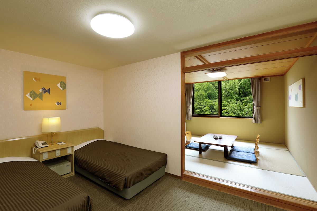 Ett hotellrum med en säng, ett nattduksbord, en lampa och utsikt över en matsal genom ett fönster.